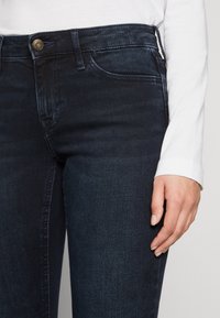 Jeans de mezclilla azul oscuro con corte slim, que cuentan con un cierre frontal de botones, presillas para cinturón y desvanecimiento mínimo. Vista en primer plano de la mano descansando sobre la cadera.
