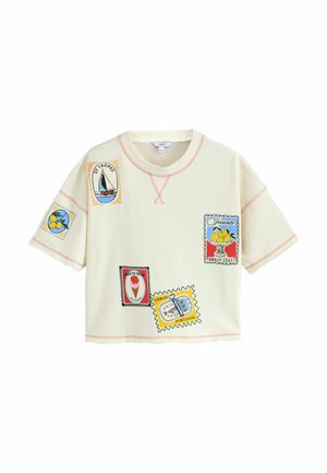 Kvinde på stranden iført beige kasket, matchende shorts og løs T-shirt med farverige rejseinspirerede patches, armbånd på venstre håndled.
