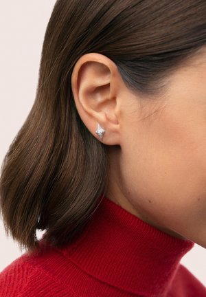 Strieborný náušník na puzetu v tvare hviezdy s textúrovaným povrchom, nosený na krátkom hnedom bob účese, na jemnom ružovom pozadí.