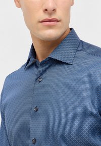 Blau gemustertes Hemd mit Knöpfen aus gewobenem Stoff, das ein kleines geometrisches Design, einen Punktkragen und dunkle Knopfakzente aufweist.