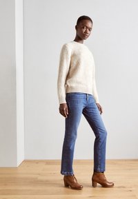Cremefarvet uldtrøje med ribbet kant, blå lige ben jeans og brune læder støvler med hæl og snørebånd. Moderne afslappet outfit på en model.