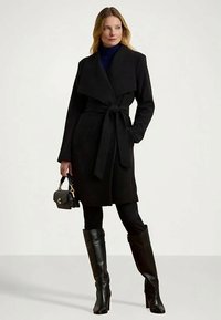 Donna che indossa un cappotto nero con cintura, leggings neri, stivali neri al ginocchio e tiene una piccola borsa nera, in piedi contro uno sfondo neutro.