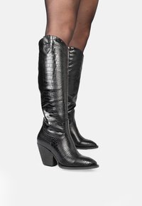 Botas de cuero negro con estampado de cocodrilo hasta la rodilla, con punta afilada y tacón grueso, que presentan un acabado brillante y detalles de costura en los laterales.