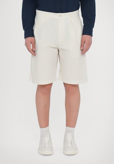 Shorts din bumbac crem cu o siluetă ajustată, prezentând pliuri și închidere cu nasture, asortați cu adidași și șosete albe.