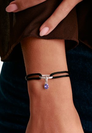 Handgelenk mit schwarzem Armband aus doppeltem Band, versehen mit silbernem Verschluss und kleinem ovalem violettem Edelsteinanhänger, braune Ärmelsaum zurückgeschoben.