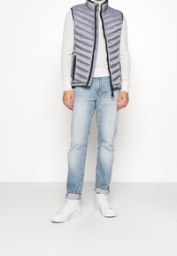 Gepolsterte graue Weste mit horizontalen Nähten, Frontreißverschluss und schwarzem Besatz. Trägt man über einem weißen Pullover und kombiniert mit hellblauen Jeans und weißen Turnschuhen.