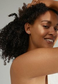 Trois petites boucles d'oreilles en perles sur l'oreille d'un modèle ; teint de peau moyen, cheveux bouclés. Maquillage léger ; épaule nue et tissu en tricot texturé visibles.