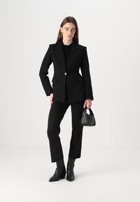 Črn tailorski blazer s enim gumbom, v kombinaciji s ožjimi črnimi hlačami. Dodatki vključujejo majhno črno torbico in čevlje s peto.