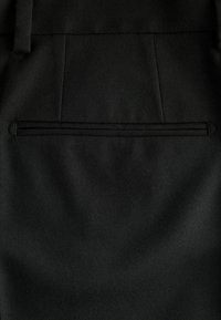 Schwarze Stoffhosen mit einer subtilen Fischgrätstruktur und einer einzelnen Gesäßtasche mit dünner, erhabener Nahtdetails.