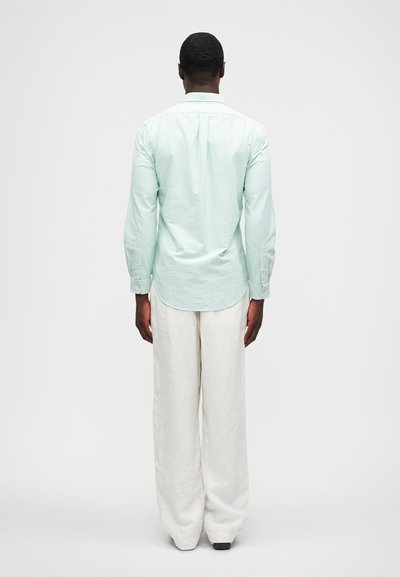 Ανοιχτό πράσινο πουκάμισο με ρίγες, μακριά μανίκια και κουμπιά στον καρπό, συνδυασμένο με χαλαρά λευκά λινά παντελόνια, παρουσιαζόμενο από πίσω.