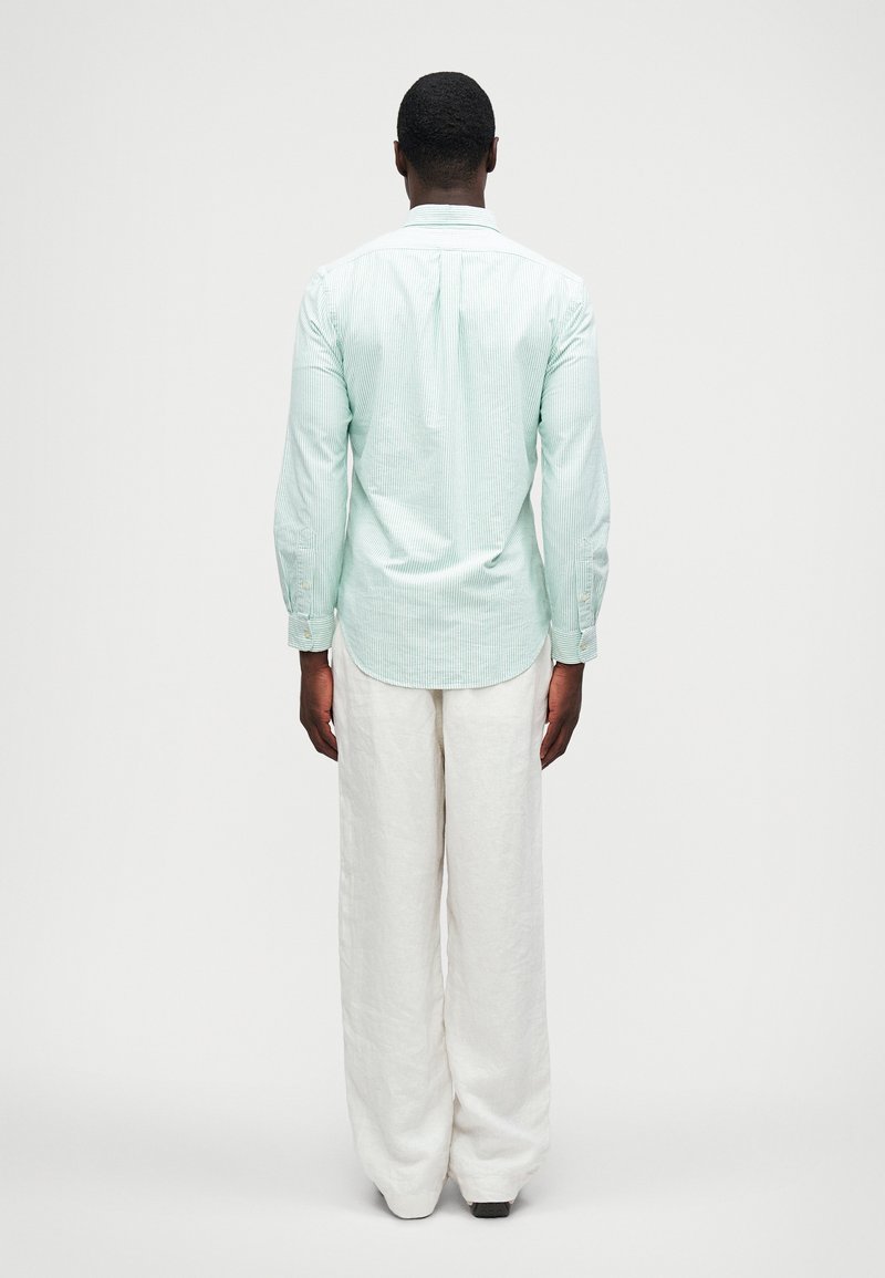 Ljusgrön randig skjorta med långa ärmar och knappmanschetter, kombinerad med löst sittande vita linnebyxor, sedd bakifrån.