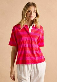 Ružovo-červený pruhovaný polo tričko s krátkym rukávom a golierom. Má uvoľnený strih a malé logo. Nosí sa s bielymi nohavicami na šnúrku.