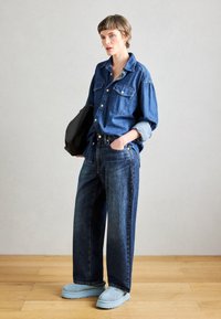 Persoon in een losse spijkerblouse en jeans, lichtblauwe instappers, met een zwarte tas in de hand, staand op een houten vloer tegen een grijze muur.