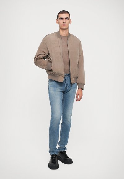Veste bomber marron clair avec des détails texturés, portée sur un haut beige et un jeans bleu, associée à des chaussures noires à lacets. Fond uni.