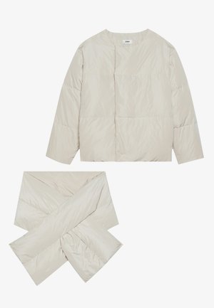 Veste beige matelassée avec un col rond, des manches longues et une écharpe assortie en forme de croix, fabriquée dans un tissu lisse et légèrement brillant.