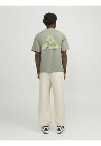 Oliven grøn t-shirt med et trekantet gult grafik på ryggen, parret med krämfarvede bukser og marineblå sneakers. Korte ærmer.
