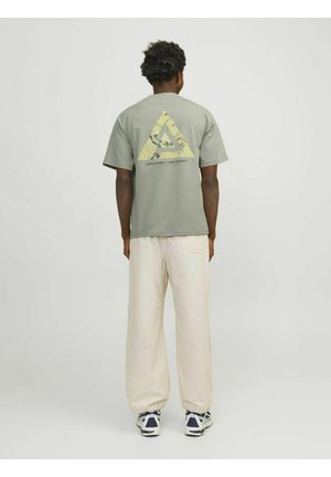Oliven grøn t-shirt med et trekantet gult grafik på ryggen, parret med krämfarvede bukser og marineblå sneakers. Korte ærmer.