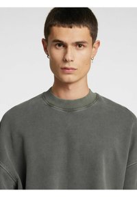 Pelēks pārizmēra džemperis ar apaļu kaklu, ribota auduma aprocēm un mīkstu tekstūru. Raksturīga brīva piegriezuma un minimālistisks dizains.