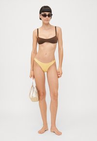 Brūns bandeau bikinis tops, apvienots ar dzelteniem bikini apakšiem, papildināts ar salmu somu un saulesbrillēm. Gluda auduma, minimāls dizains.