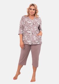 Kobieta stoi boso, ubrana w fioletową, kwiatową koszulę zapinaną na guziki z rękawami do łokci oraz dopasowane spodnie typu cropped, trzymając jedną rękę w kieszeni.