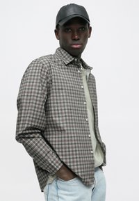 Camicia a quadri con bottoni in toni grigio e marrone smorzati, realizzata in tessuto leggero. Abbinata a una canotta verde chiaro e un berretto nero.