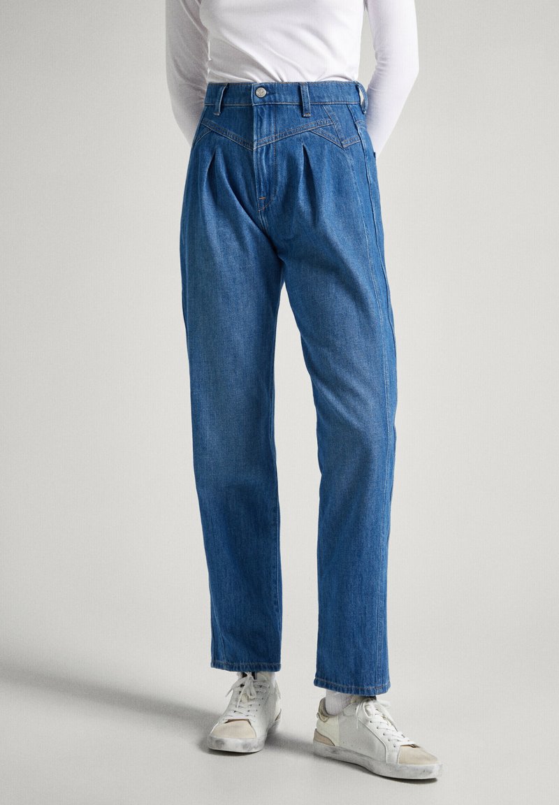 Pantalones de mezclilla azules con una cintura alta, pliegues en la parte frontal y un diseño de pierna ajustada. Combinados con zapatillas blancas.