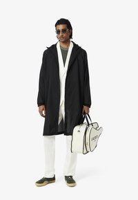 Zwarte lange regenjas, witte blazer, groene T-shirt, witte broek, groene sneakers. Houdt een witte Lacoste tas vast met zwarte accenten en details.