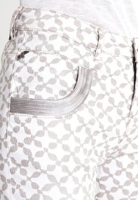 Jeans blancos con un patrón de hojas beige, con un brillante acento plateado en el bolsillo delantero. Hechos de tela de mezclilla con una textura suave.