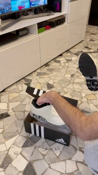 Balta Adidas sporta apavu ar auduma augšdaļu un melnām detaļām. Rokā tiek turēta kurpe no melnas kastes ar baltām svītrām, kas atrodas uz flīzēta grīdas seguma.