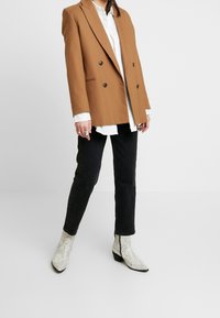 Blazer marron croisé porté sur une chemise blanche, associé à un jean noir et des bottines blanches à imprimé serpent, avec des bouts pointus et des talons bas.