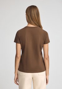 Camiseta corta marrón de manga corta en tejido suave, con un clásico cuello redondo y una silueta ajustada, combinada con pantalones de color claro.