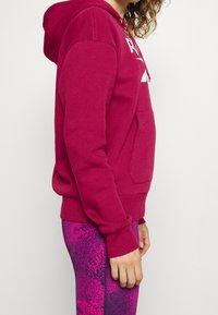 Fuchsia huvtröja med känguruficka, ribbstickade ärmslut och dragsko i huven, bärs med mönstrade rosa leggings.