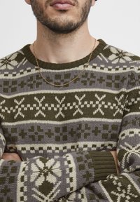 Strickpullover mit geometrischem Muster in Olivgrün, Creme und Grau, mit gerippten Bündchen und rundem Halsausschnitt. Enthält eine dünne goldene Halskette.