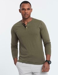 Man i olivgrön långärmad henley-tröja med tre knappar, vita byxor och svart armbandsur, stående med en hand i fickan.