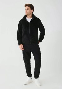 Férfi fekete bolyhos, cipzáras dzsekiben, fekete katonai nadrágban és fehér sportcipőben, az egyik kezét zsebre dugva, oldalra nézve áll.