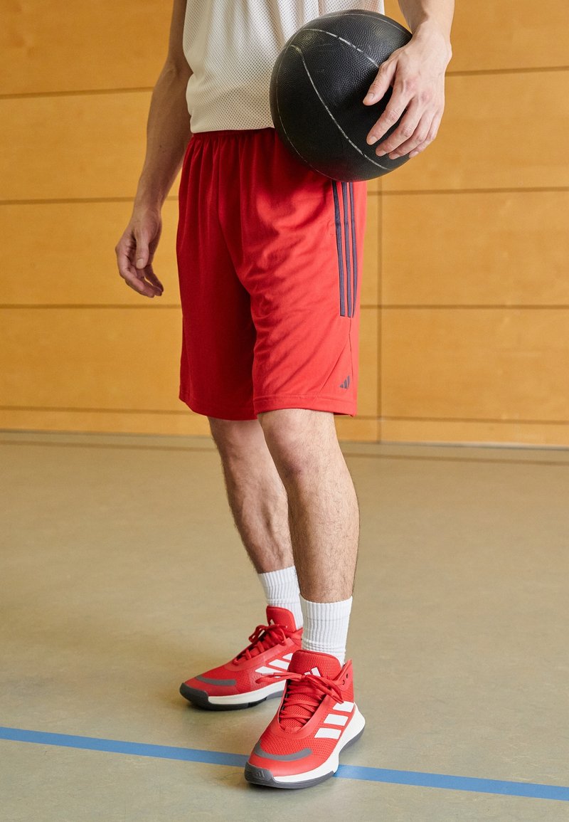 Personne en short de sport rouge et chaussures tenant un ballon de basket noir, debout sur un terrain intérieur avec un mur en bois en arrière-plan.