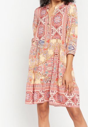 Vrouw die een knielange jurk met lange mouwen draagt met ingewikkelde rood, roze en beige bloemen- en geometrische patronen, staand met één hand op haar dij.