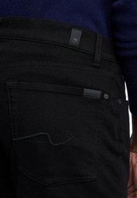 Zwarte denim jeans met een platte achterzak, subtiele stiksels en een klein merketiket nabij de tailleband, getoond vanuit de achterkant.