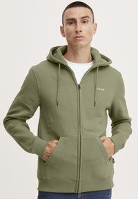 Felpa con cappuccio e zip in tessuto verde oliva, dotata di tasche a marsupio, cappuccio con coulisse e logo stampato sul petto.