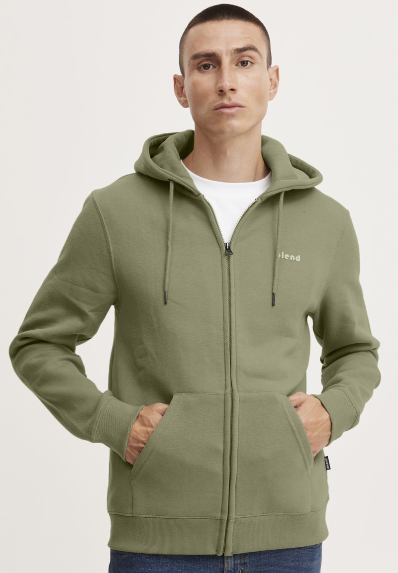 Felpa con cappuccio e zip in tessuto verde oliva, dotata di tasche a marsupio, cappuccio con coulisse e logo stampato sul petto.