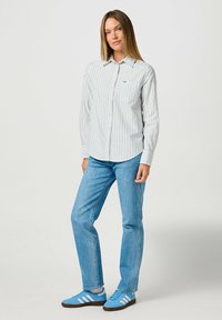 Camicia bianca con righe verticali blu, abbinata a jeans azzurri chiari e sneakers blu con righe bianche. Taschino sul petto ben visibile.