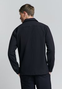 Zwarte softshell jack, lange mouwen, puntige kraag, subtiele naaddetails en knoopaccenten bij de manchetten. Gladde, licht rekbare stof.