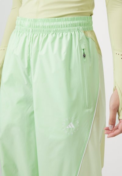 Pantalons coupe-vent brillants, vert clair, avec une taille à cordon de serrage, une poche zippée sur le côté et un logo blanc près de l'ourlet. Texture lisse.