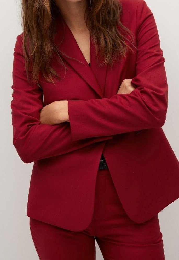 Femme aux longs cheveux bruns portant une veste de costume rouge et un pantalon assorti, les bras croisés devant un fond clair uni.