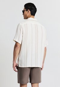 Hemd mit kurzem Arm und Kragen in hellem Creme mit horizontalen Streifen; strukturierter Stoff, kombiniert mit braunen Shorts. Figurbetontes, lässiges Design.