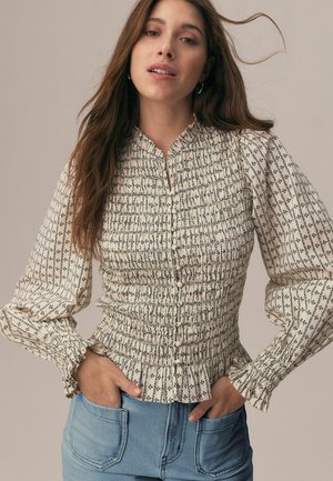 Młoda kobieta o długich brązowych włosach, ubrana w kremową, kwiecistą bluzkę z marszczeniami i jasnoniebieskie dżinsy, z rękami w kieszeniach, na jednolitym tle.