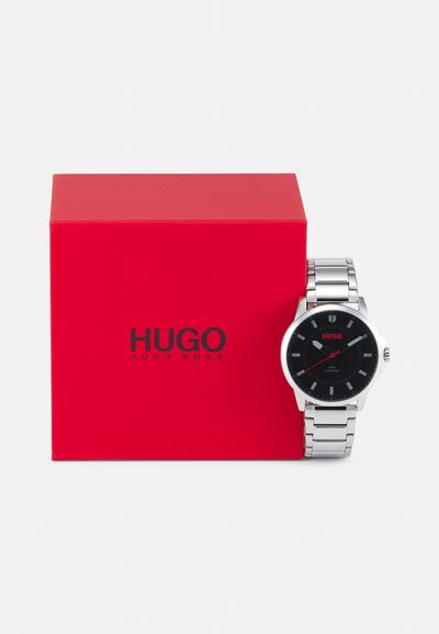 Montre en métal argenté avec un cadran noir, agrémentée d'accents rouges et de repères horaires, à côté d'une boîte rouge arborant le logo "HUGO".