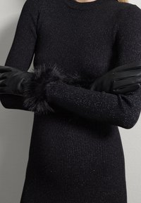 Jersey negro de canalé con hilos metálicos, que presenta mangas largas y un puño de piel sintética negra, combinado con unos elegantes guantes de cuero negro.