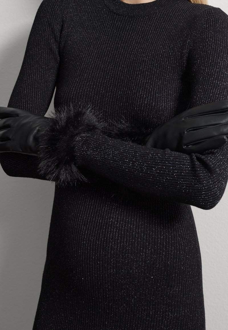 Jersey negro de canalé con hilos metálicos, que presenta mangas largas y un puño de piel sintética negra, combinado con unos elegantes guantes de cuero negro.
