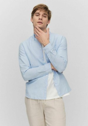 Chemise boutonnée bleu clair à manches longues, fabriquée en tissu léger, avec une coupe décontractée et un léger éclat. Portée sur un t-shirt blanc.
